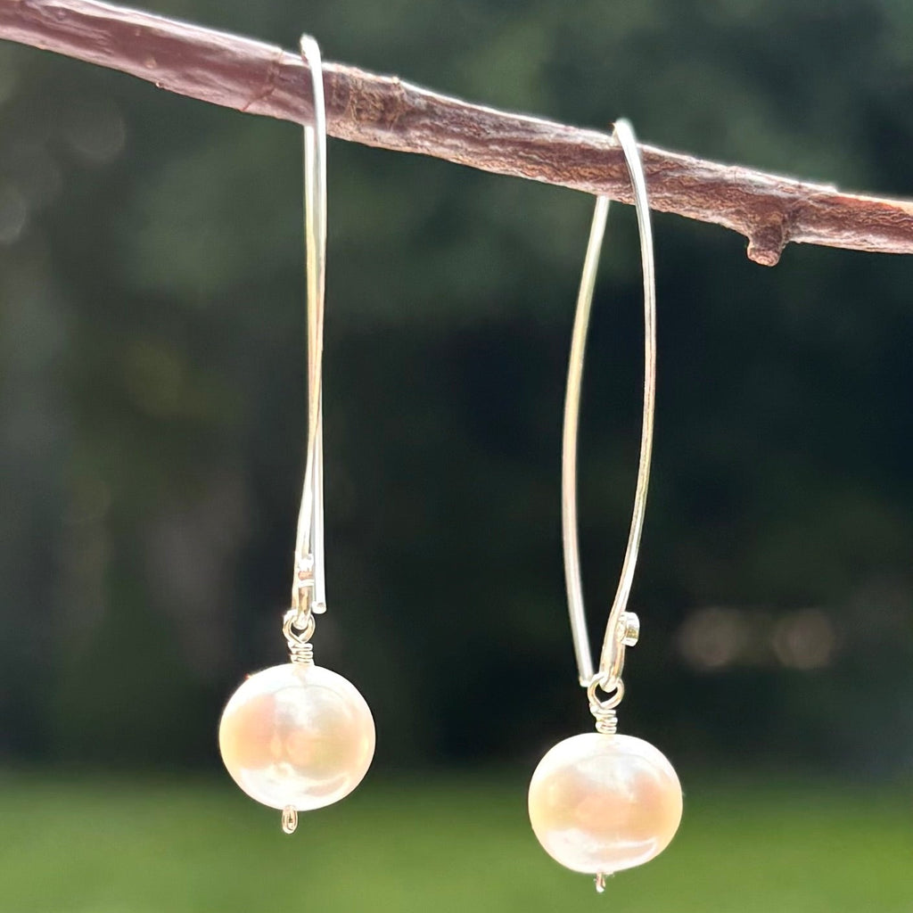 A pair of silver hoop earrings with round, pale pinkish-beige pearl-like drops, hanging from a brown branch against a blurred green outdoor background.