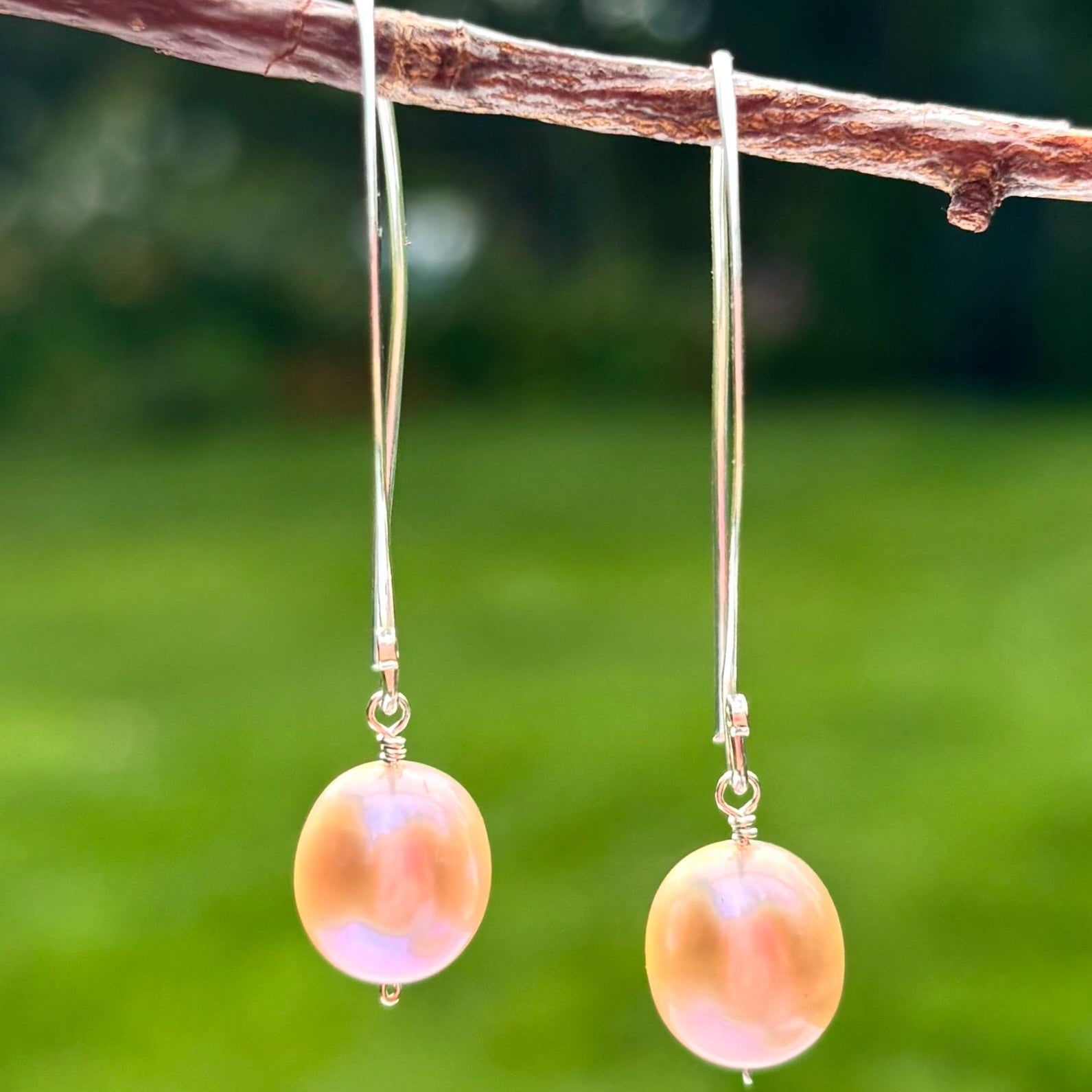 A pair of dangling earrings with round iridescent pink pearls on silver hooks, hanging from a brown branch against a blurred green outdoor background.
