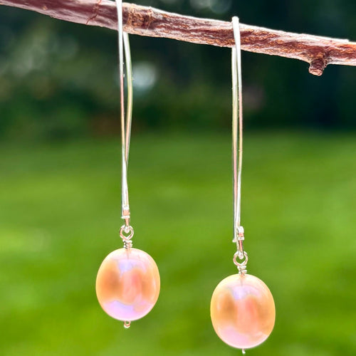 A pair of dangling earrings with round iridescent pink pearls on silver hooks, hanging from a brown branch against a blurred green outdoor background.