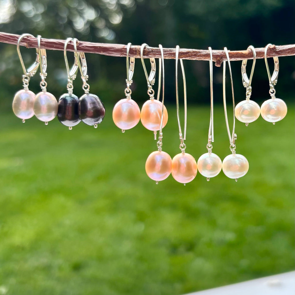 A set of dangling earrings hanging from a thin branch outdoors, with a blurred green lawn and trees in the background. The earrings feature round pearl-like beads in peach, cream, and dark brown/black, suspended from silver hooks.