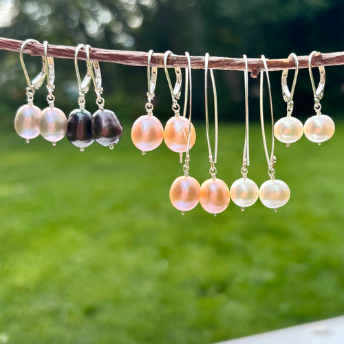 A set of dangling earrings hanging from a thin branch outdoors, with a blurred green lawn and trees in the background. The earrings feature round pearl-like beads in peach, cream, and dark brown/black, suspended from silver hooks.