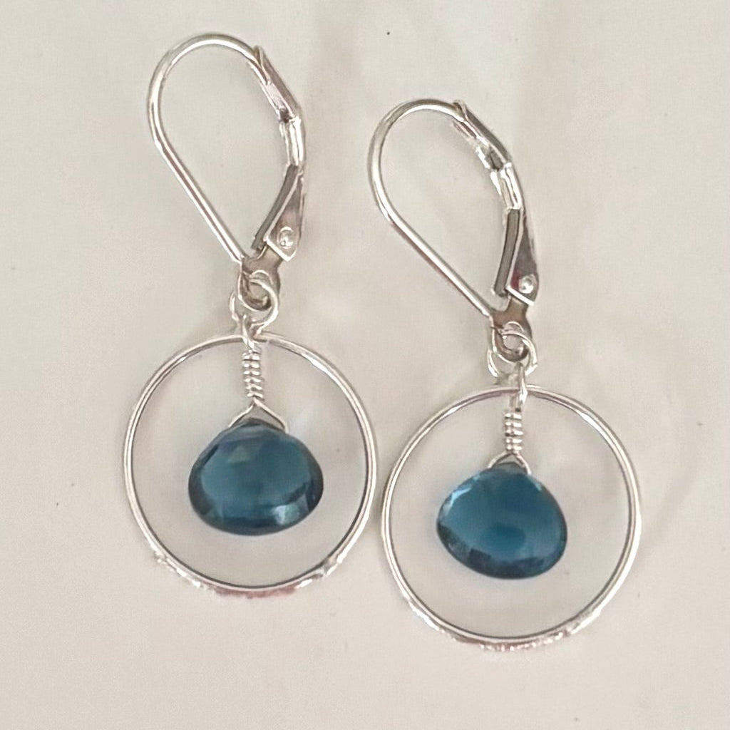 A pair of silver leverback dangle earrings with circular wire hoops and faceted blue gemstone drops, shown on a plain light background.