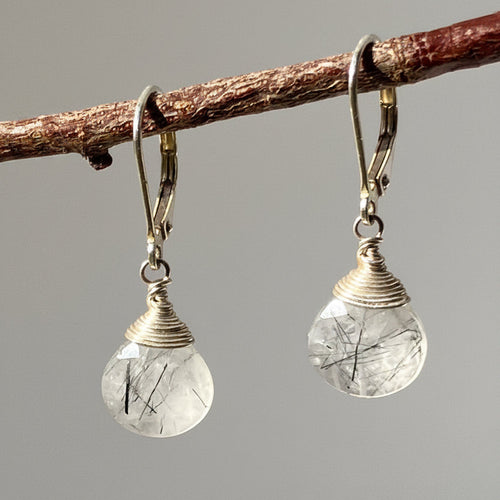 A pair of dangling earrings with clear teardrop-shaped rutilated gemstone drops and silver-toned wire-wrapped caps on silver fishhook ear wires, hanging from a brown twig against a neutral gray background.