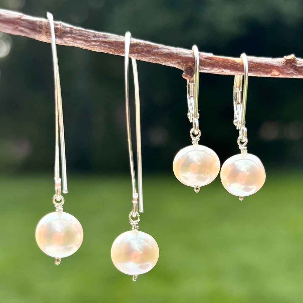 Three pairs of silver drop earrings with large iridescent pearl-like beads hanging from a natural branch, set against a blurred green outdoor background.