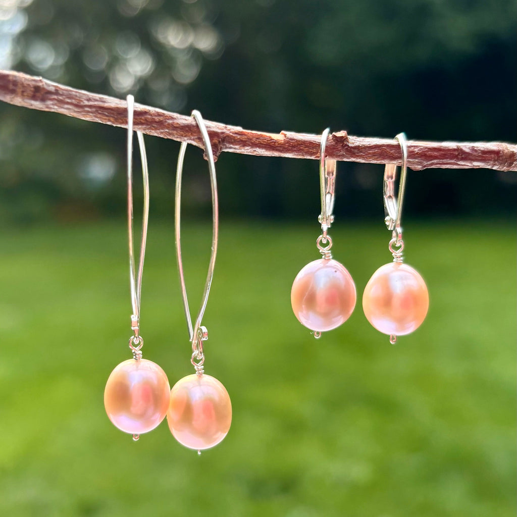 Three pairs of dangling earrings with round pink freshwater pearls and silver hooks, hanging from a brown branch against a blurred green outdoor background.