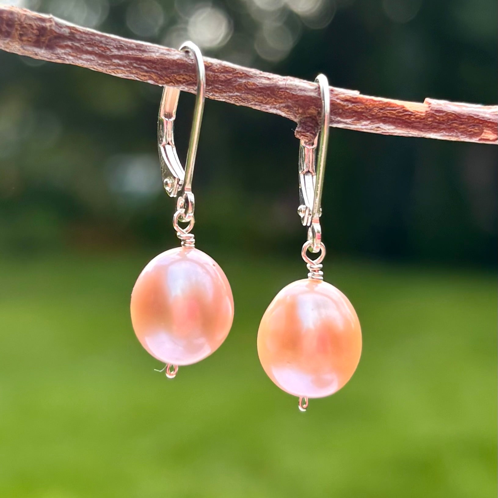 A pair of dangling earrings with large round pink baroque pearls suspended from silver fishhook wires, hanging from a brown branch against a blurred green outdoor background.